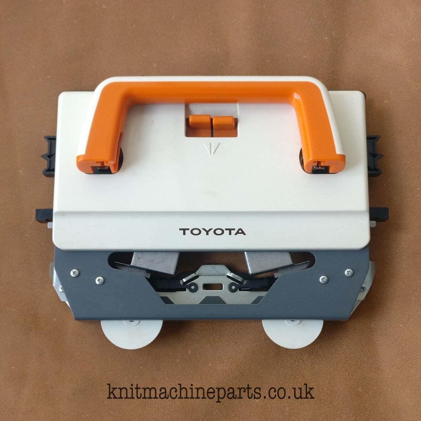 A Toyota KS 901 knitting machine lace carriage, featuring a white body with orange handles and gray accents, placed on a brown surface.