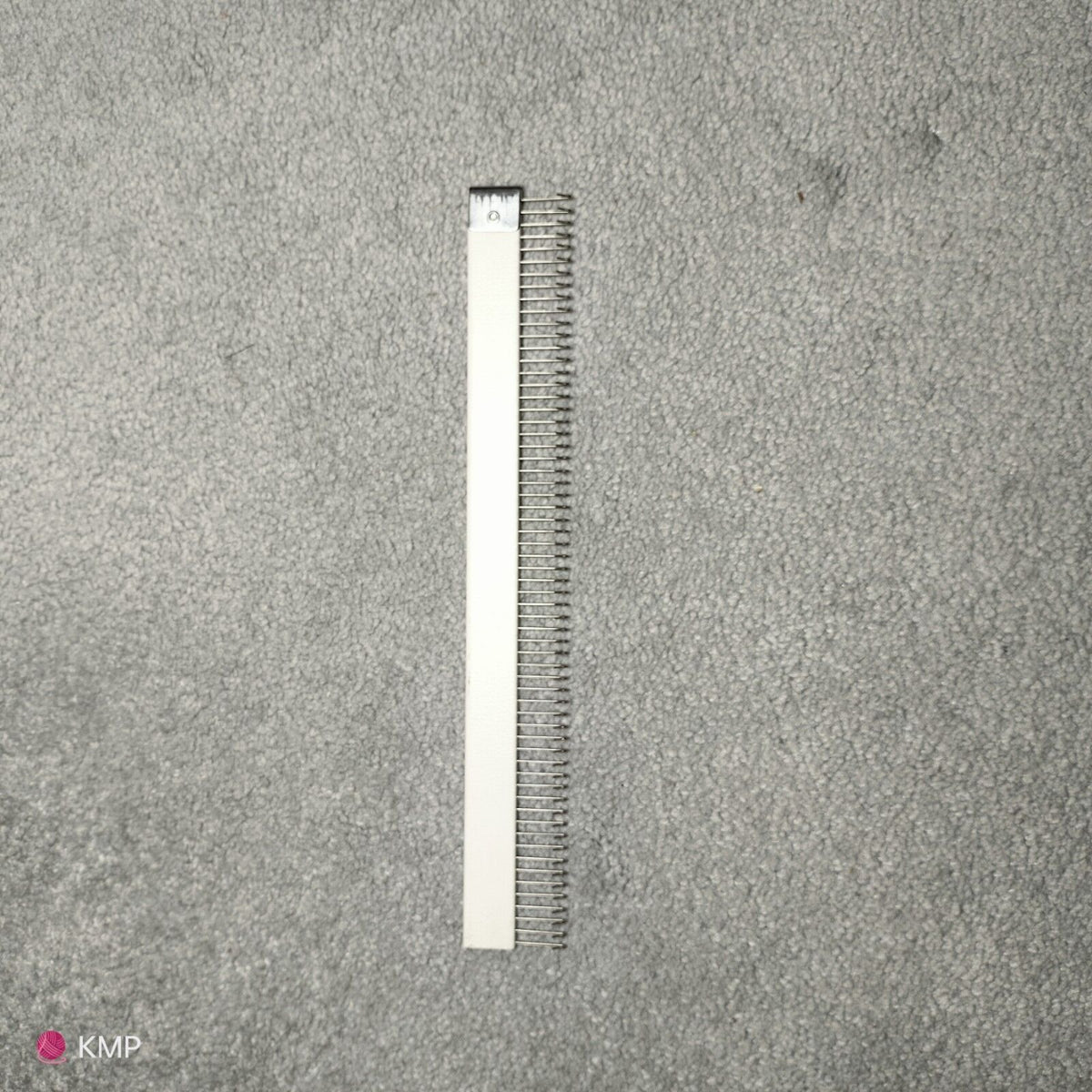 A metal cast-on comb for an electronic knitting machine Brother KH 900, lying on a grey surface.